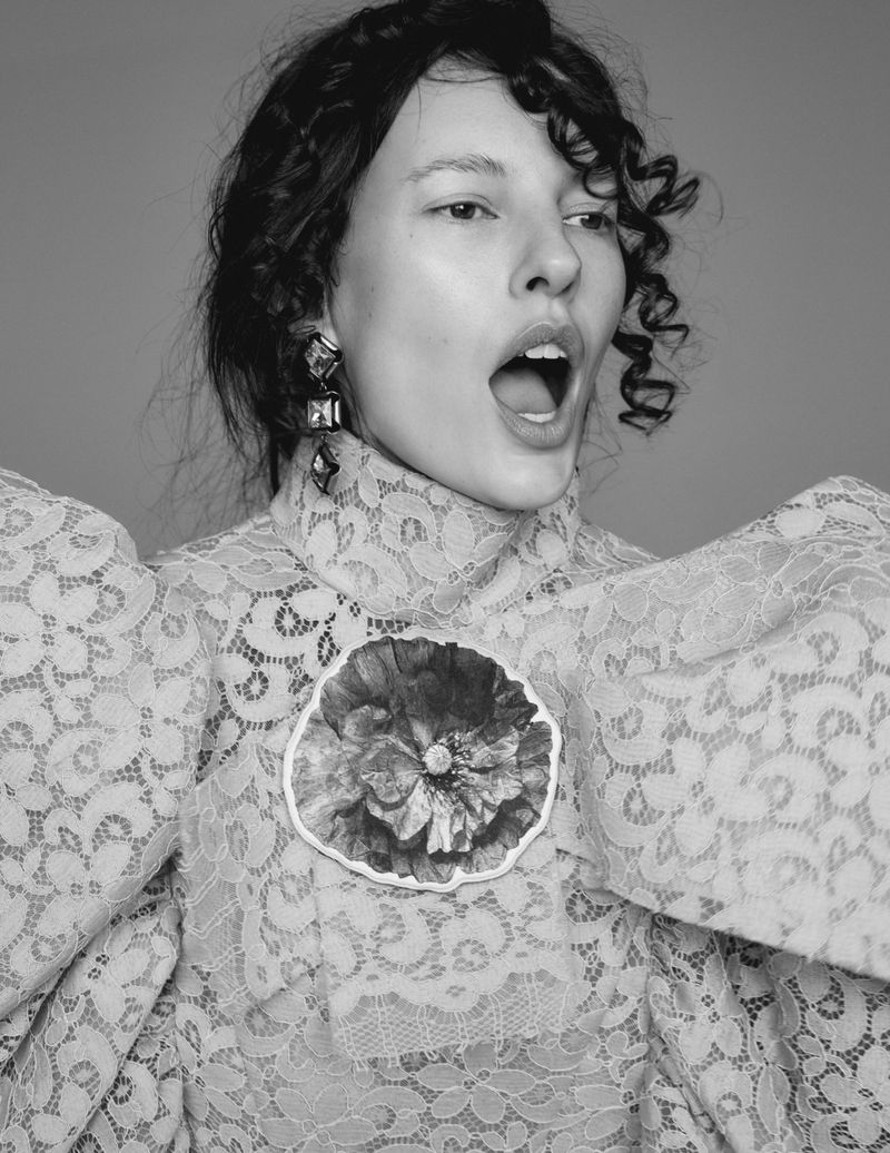 Black and white portrait of an adult female in a lace dress with a flower brooch, posing with an open mouth against a grey background.