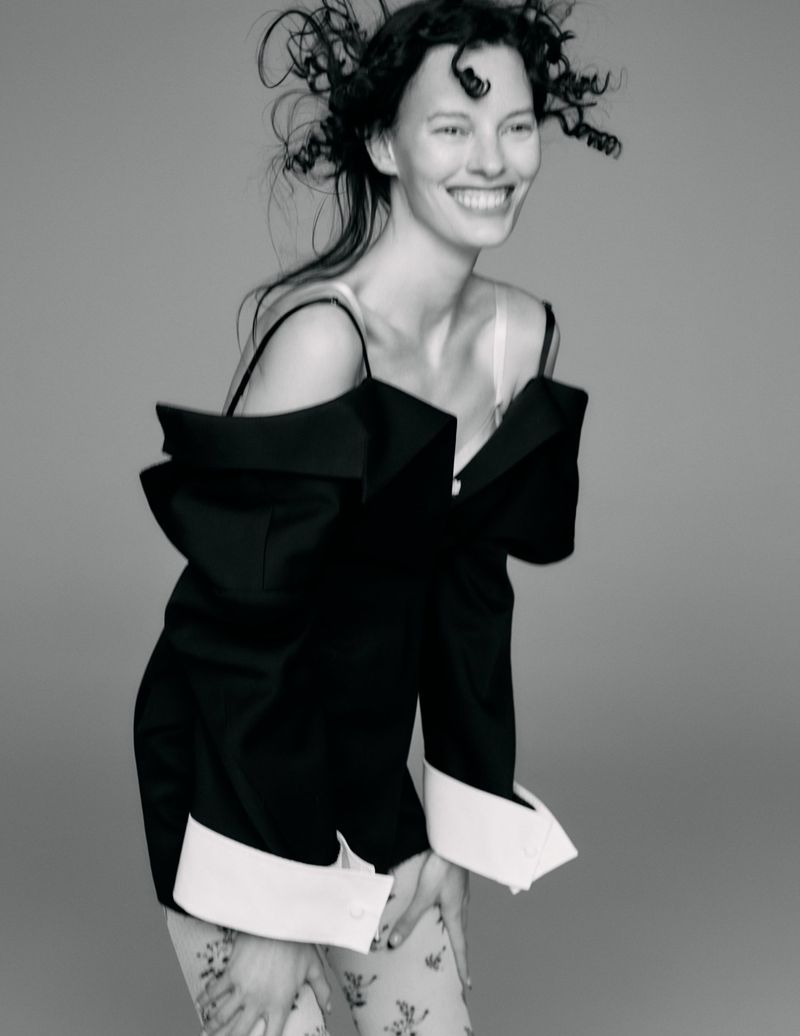 Black and white portrait of a smiling woman with curly hair wearing a black jacket and floral-patterned pants, set against a grey background.