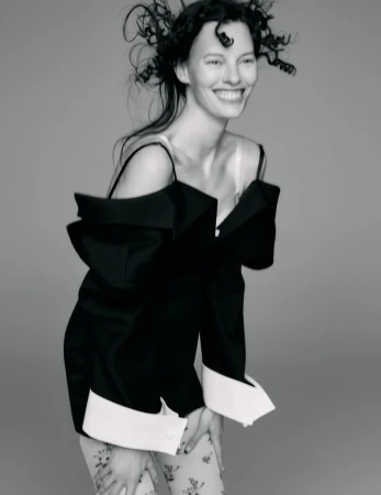 Black and white portrait of a smiling woman with curly hair wearing a black jacket and floral-patterned pants, set against a grey background.