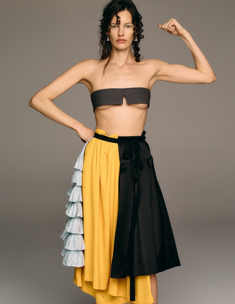 A model poses in a strapless top and color-blocked skirt in a studio setting. The model has dark curly hair and is flexing her arm.