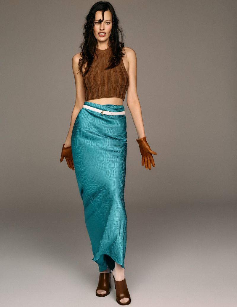 Full-length shot of a woman in a brown knit halter top, teal skirt with a white belt, and brown gloves and heels, posing in a studio.