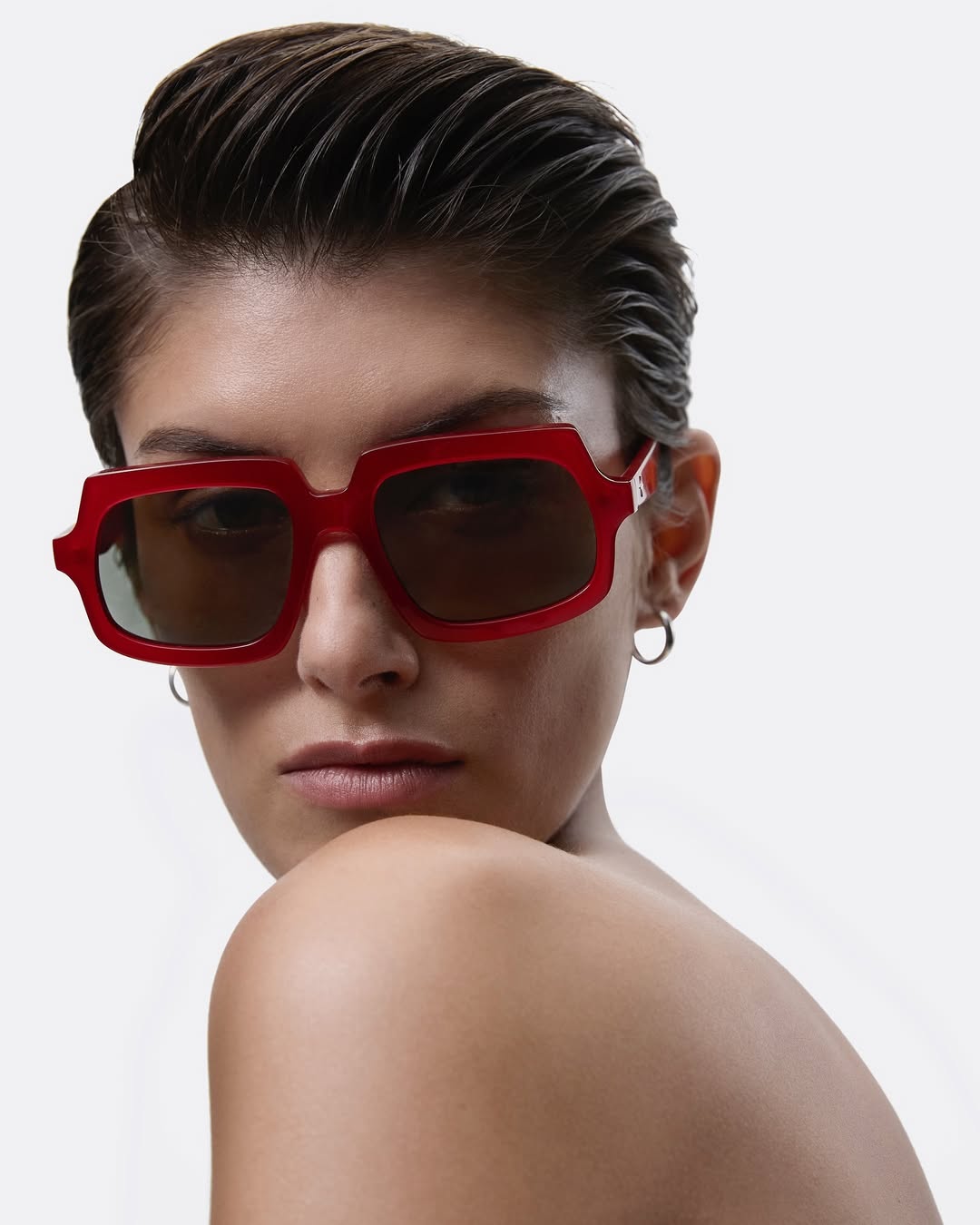 Close-up portrait of a woman wearing glossy red frame sunglasses with dark tinted lenses against a plain white background