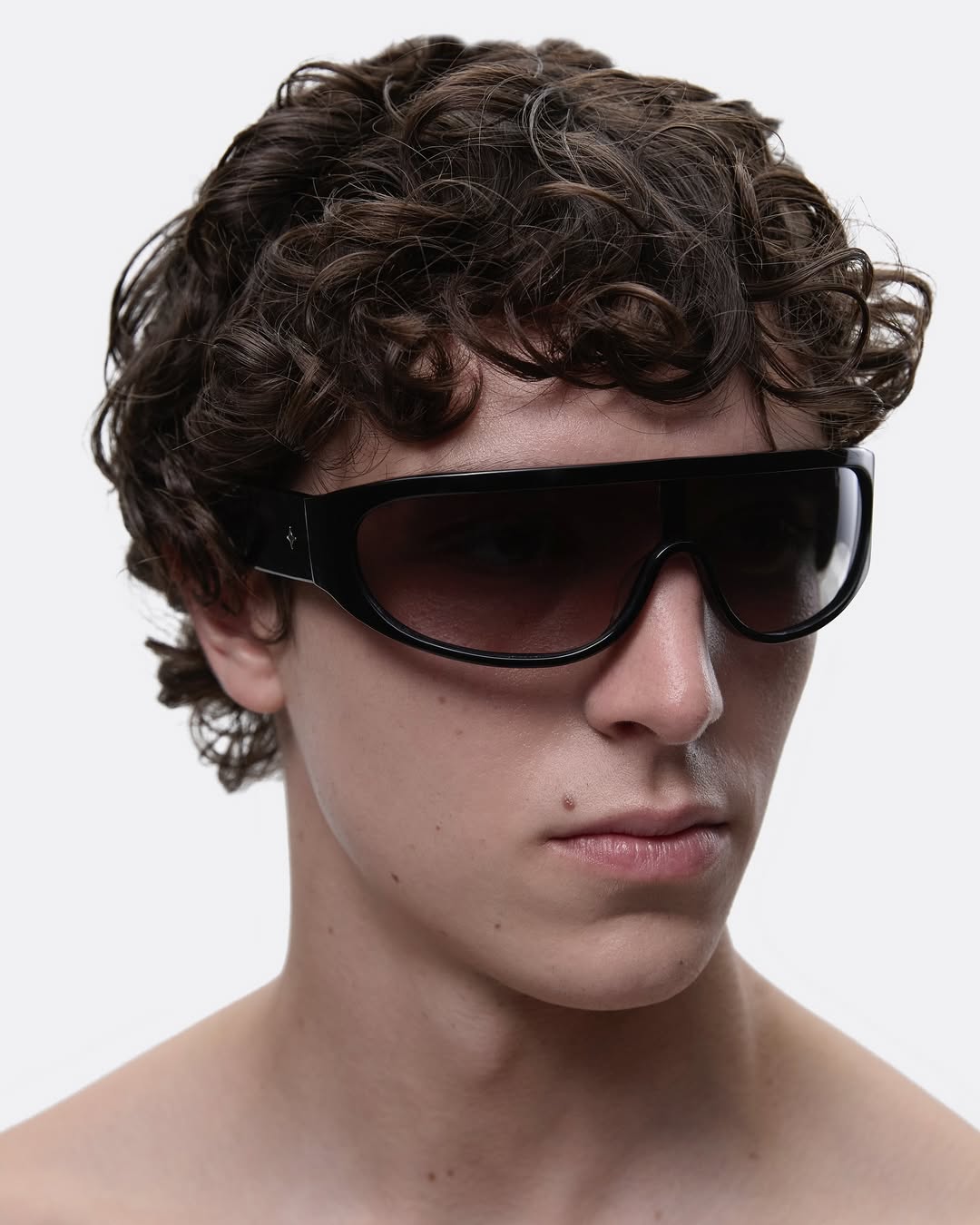 Close-up shot of a male with dark, curly hair wearing wide, black sunglasses with gradient lenses against a bright white backdrop.