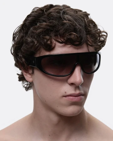Close-up shot of a male with dark, curly hair wearing wide, black sunglasses with gradient lenses against a bright white backdrop.