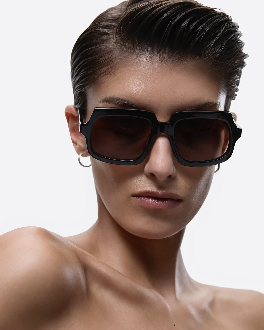 Close-up of a woman with short, dark hair wearing black sunglasses against a white background, showcasing fashion and beauty elements.