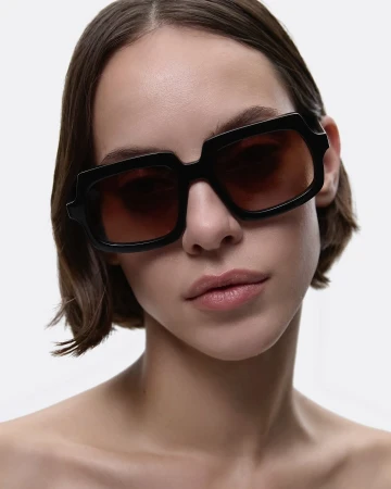 Close-up of a young adult female model in a studio wearing chunky black sunglasses with brown lenses against a white backdrop.