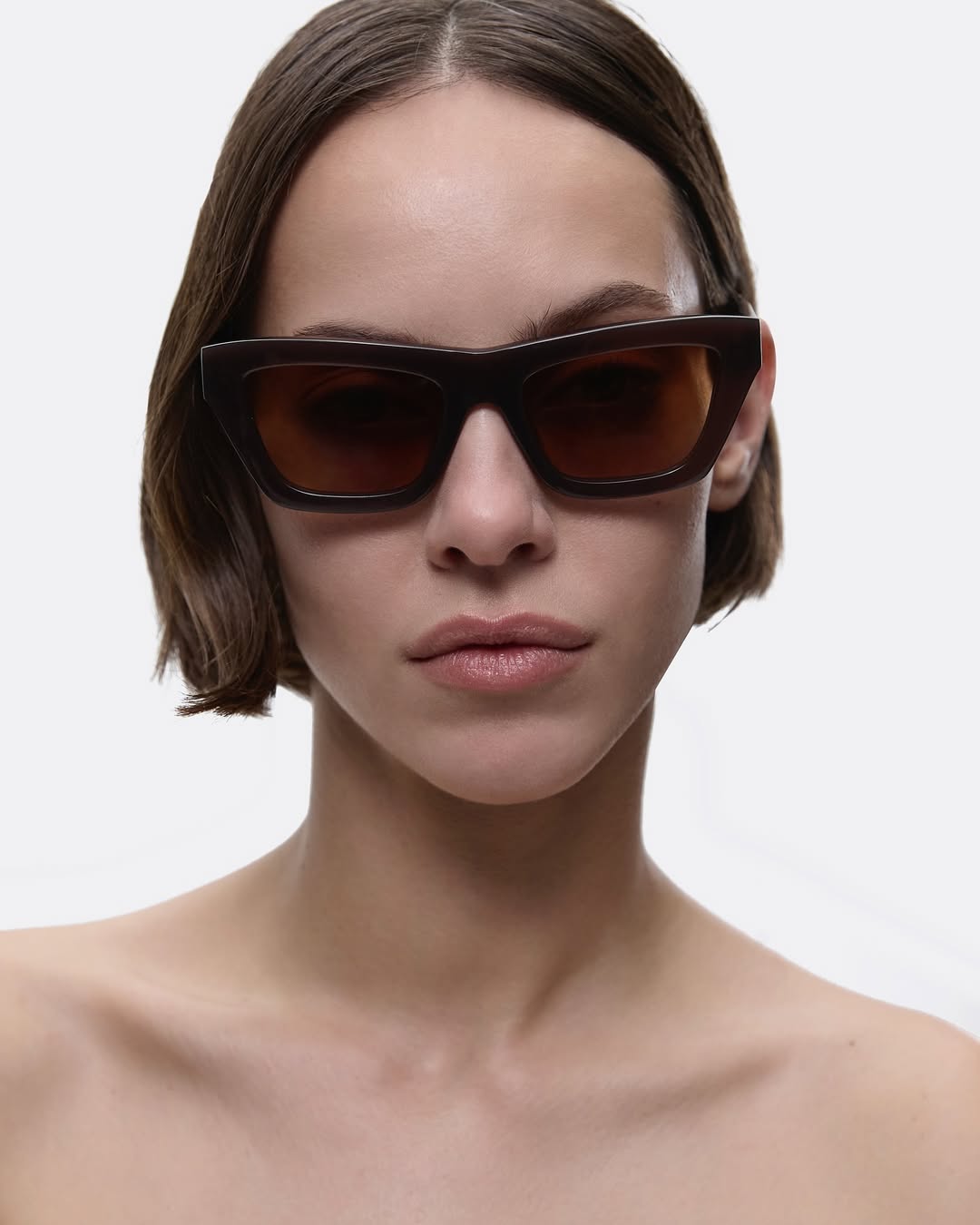 Close-up studio shot of a young adult female model wearing dark sunglasses against a bright white background. Fashion and beauty shot.