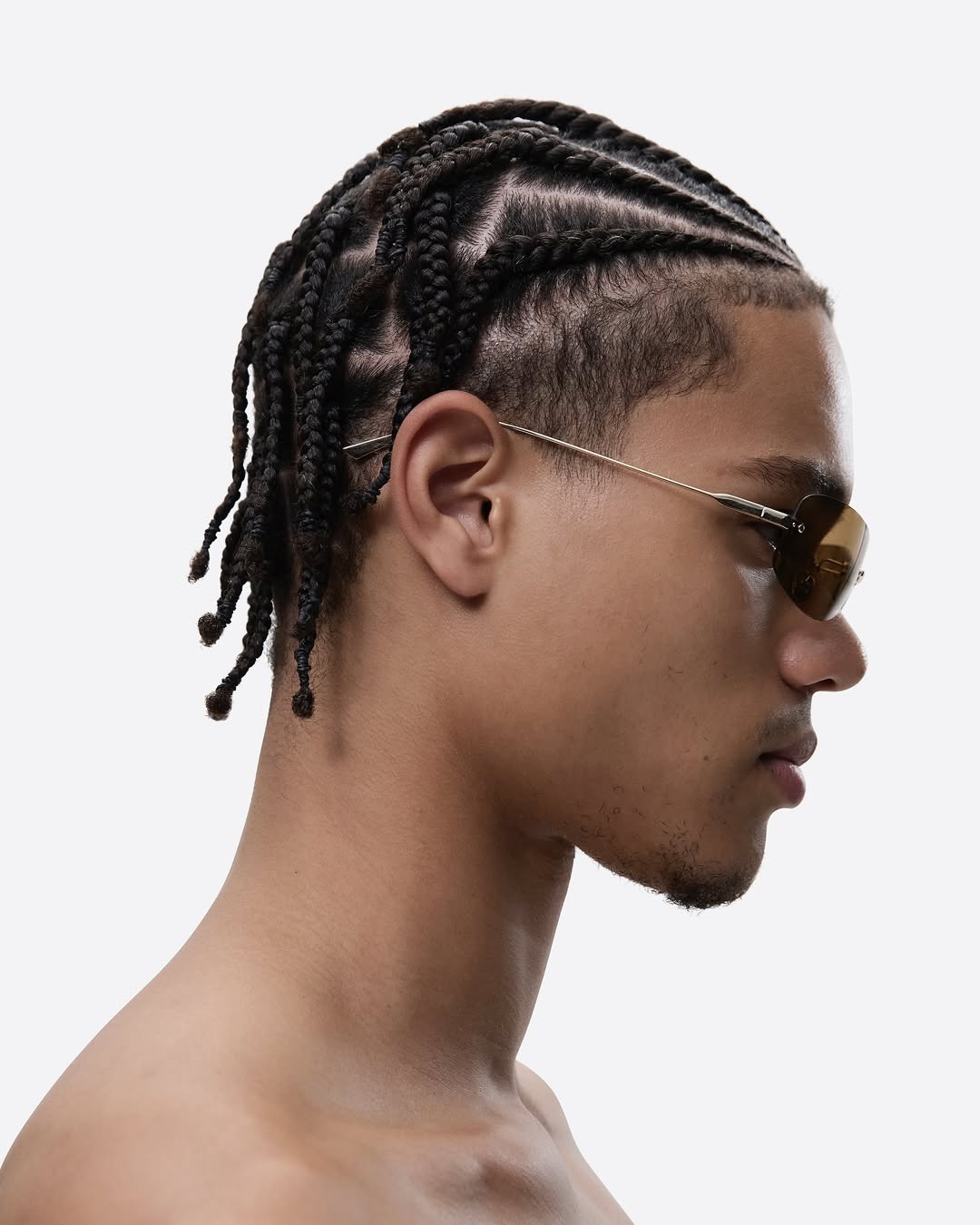 Profile view of adult male with dark braided hair and sunglasses, on a light background in a studio setting. Minimalist fashion shot.