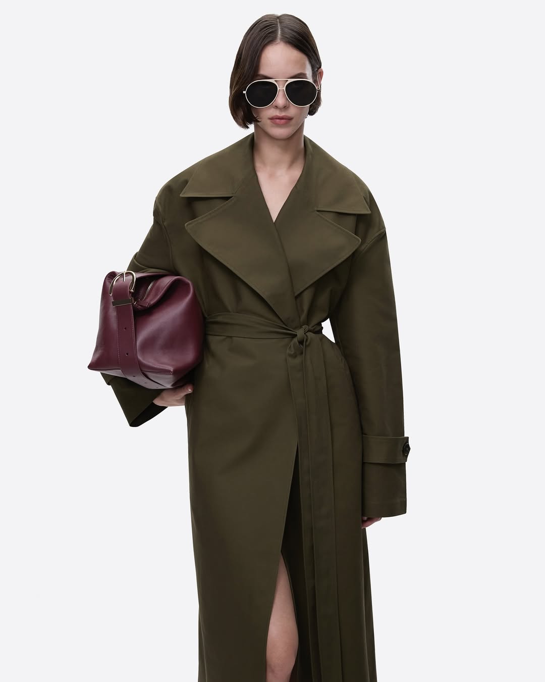 Elegant woman wearing a green trench coat and sunglasses holding a burgundy leather handbag in a studio shot against a white backdrop.