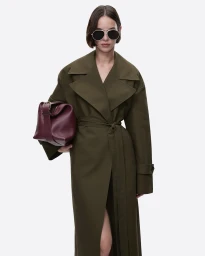 Model wearing dark sunglasses and an olive green coat holding a burgundy handbag against a white seamless background