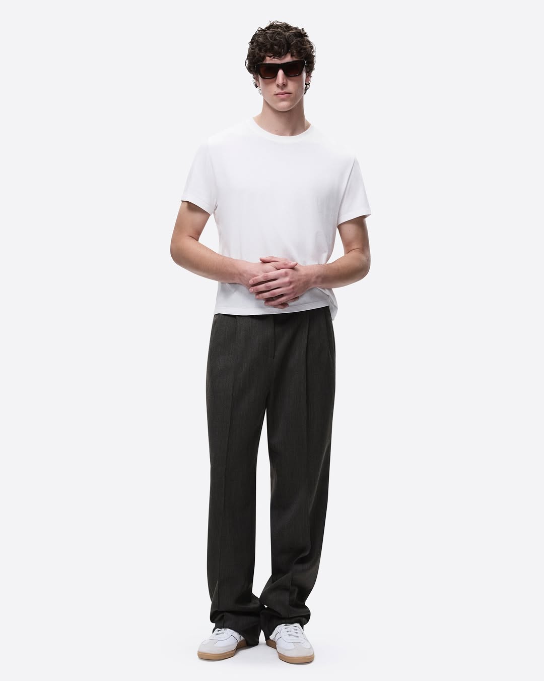 Full-length shot of a young adult male model wearing a white t-shirt, dark pants, sunglasses, and sneakers against a white background.