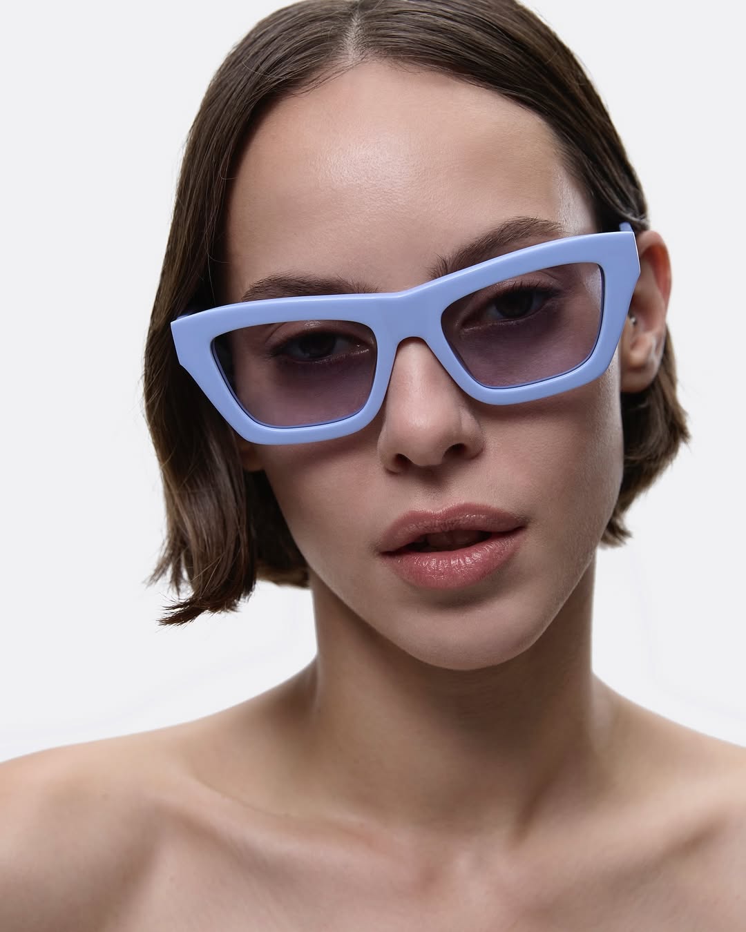 Close-up studio shot of a young woman with fair skin wearing light-blue-frame sunglasses, showcasing a modern fashion eyewear style.