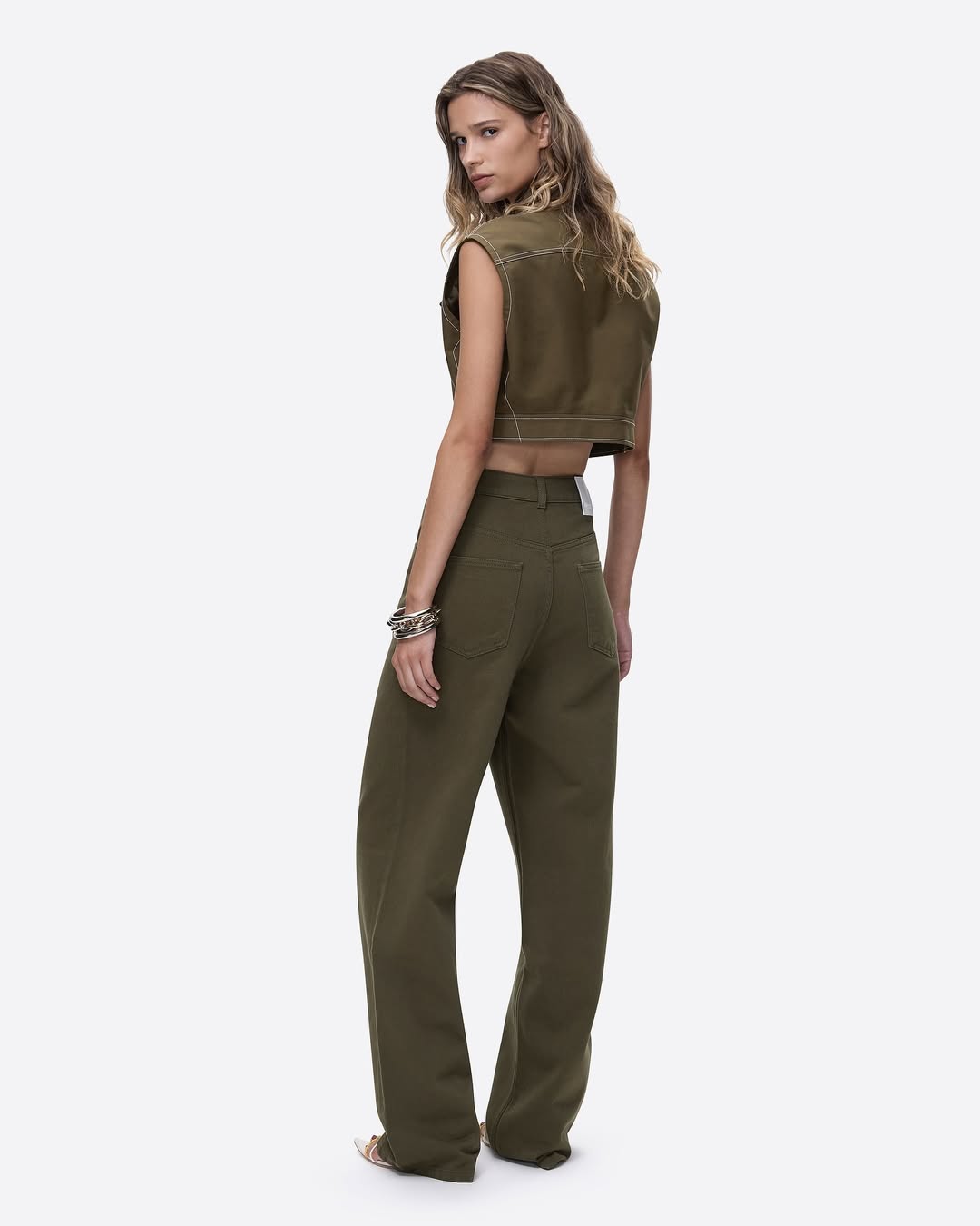 Olive green wide-leg jeans and a cropped vest worn by a model with long brown hair in a fashion studio shot on a white background.