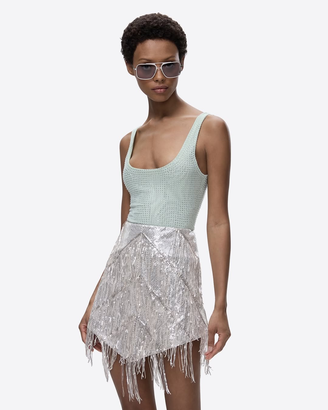 Woman poses in a light green bodysuit and silver fringed skirt against a bright background, showcasing a fashion editorial style.
