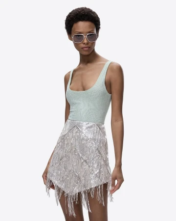 Woman poses in a light green bodysuit and silver fringed skirt against a bright background, showcasing a fashion editorial style.