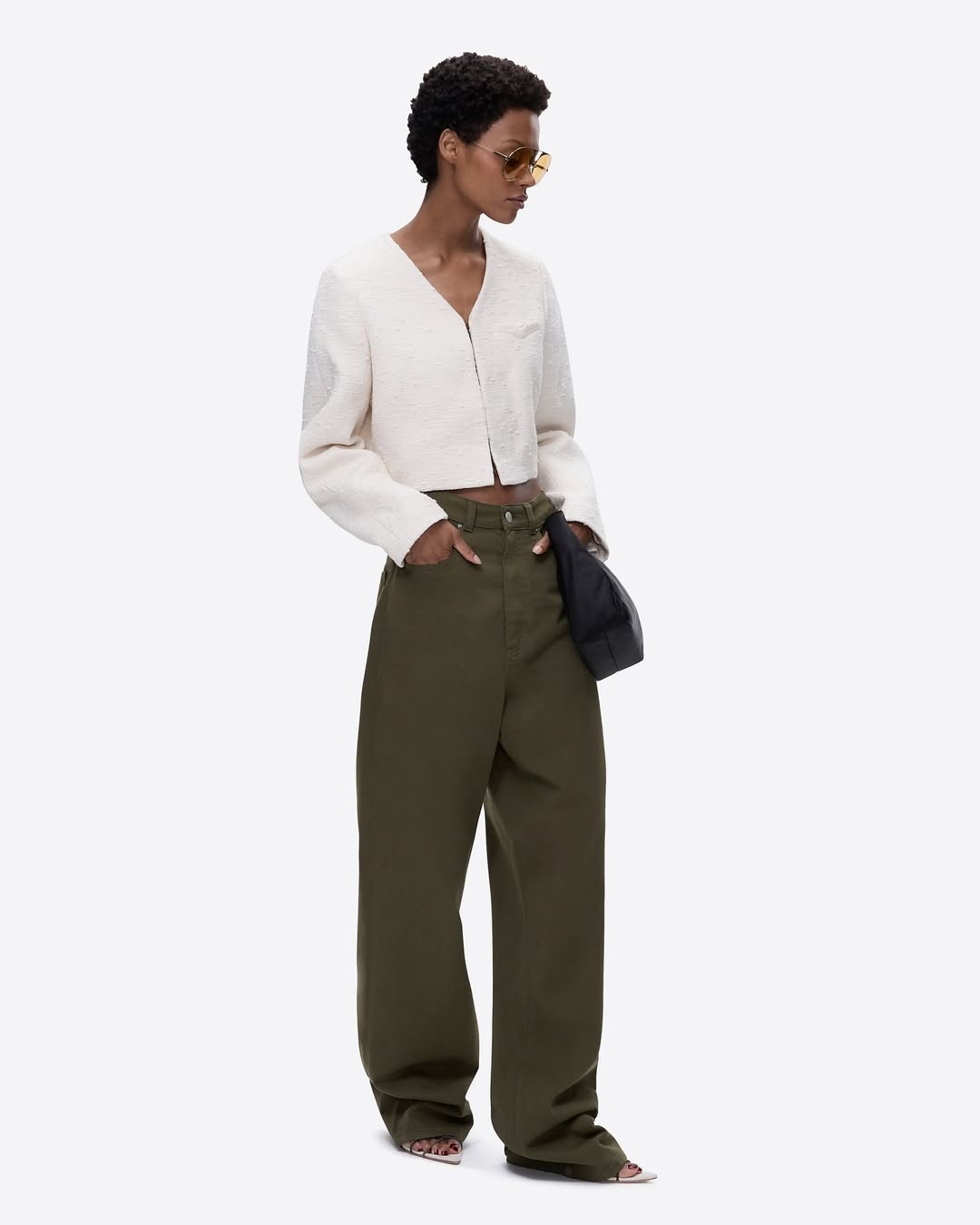 A woman in a cream jacket and olive trousers stands against a plain white background in a fashion editorial shot.