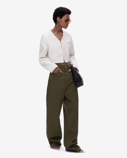 A woman in a cream jacket and olive trousers stands against a plain white background in a fashion editorial shot.