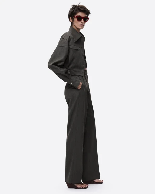 An adult woman with short hair poses in a grey-green collared crop jacket, high-waisted palazzo pants, and burgundy sunglasses against white.
