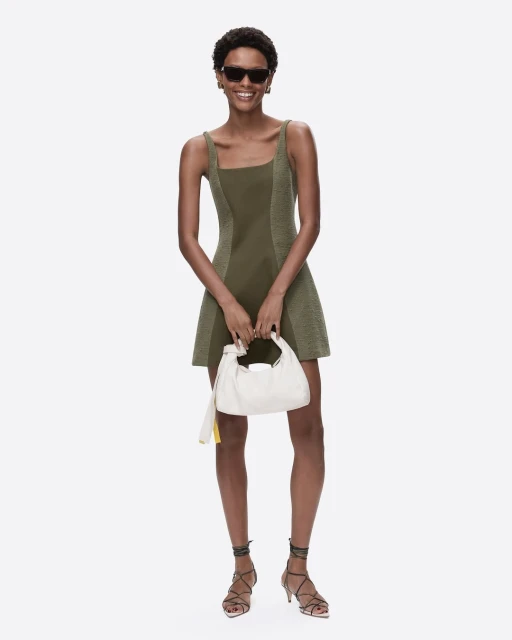 A cheerful adult woman smiles while wearing an olive-green mini dress, strappy heels, and sunglasses, and holding a white purse.