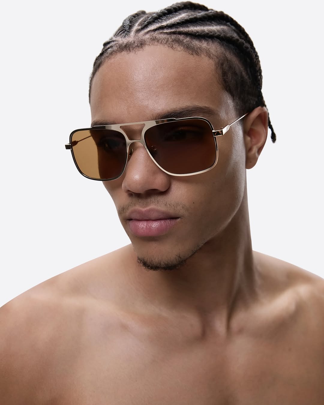A close-up studio shot of an adult male model with cornrows, wearing gold-rimmed aviator sunglasses against a white background.