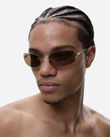 Close-up studio shot of a young adult male wearing rimless tinted sunglasses. He has braided hair and is partially nude on a white backdrop.