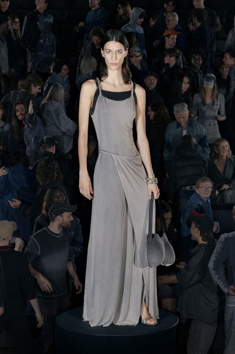 Fashion show featuring a model wearing a long grey dress with a high slit, accessorized with a bracelet and matching grey purse, standing before an audience.