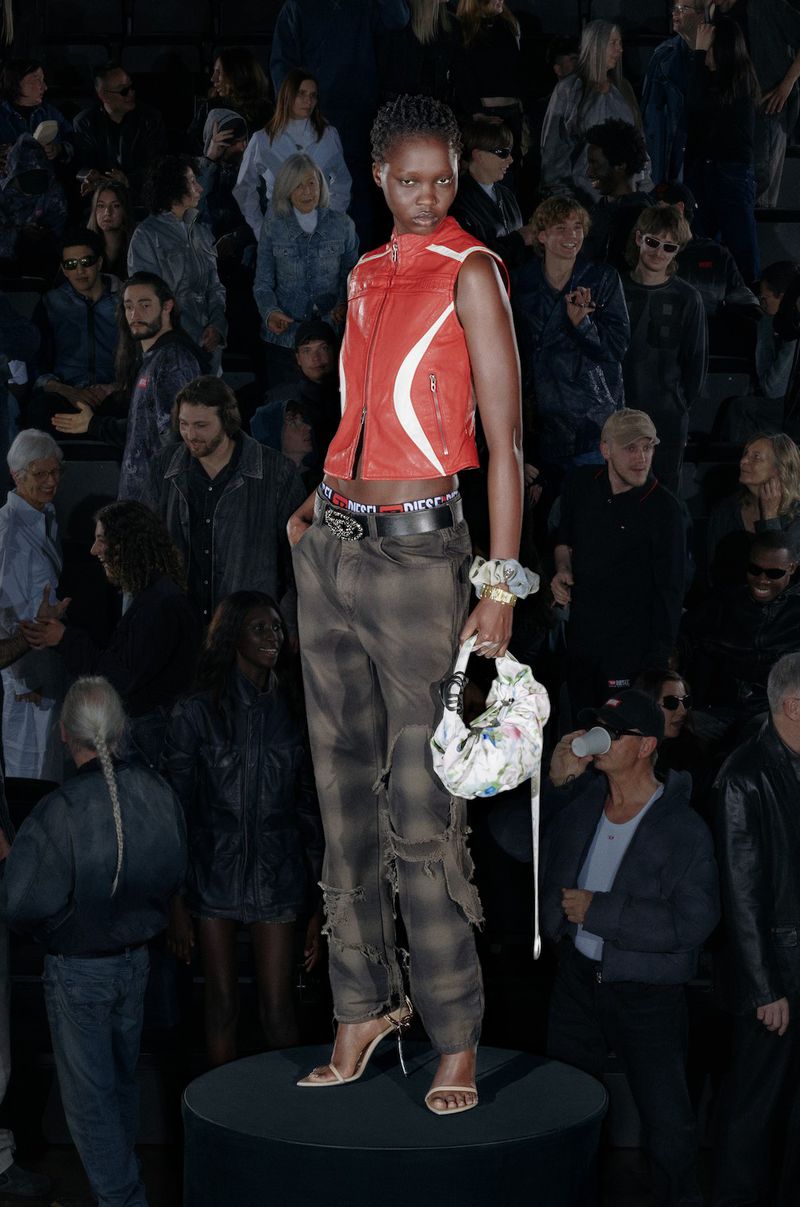 Runway fashion: a model stands on a black platform, wearing a red vest, patterned pants, and high heels, surrounded by a dark audience.