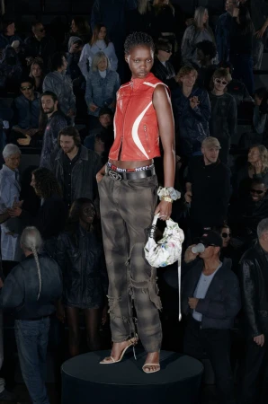 Runway fashion: a model stands on a black platform, wearing a red vest, patterned pants, and high heels, surrounded by a dark audience.