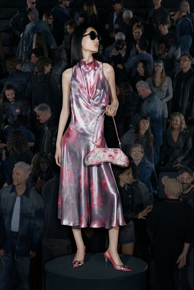 Asian model poses with a floral purse wearing a shiny pink gown, standing on a dark platform with a crowd of people in the background.