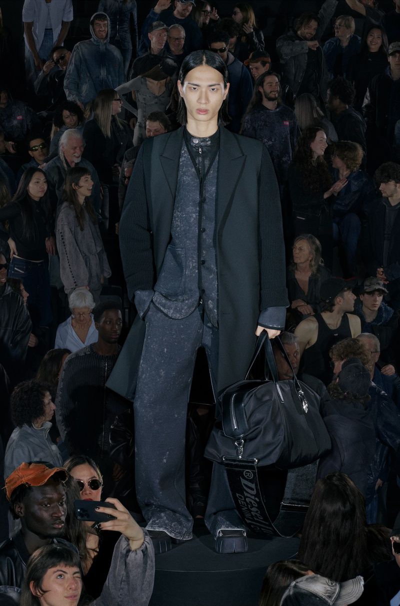 Asian male model on runway wearing dark grey suit, vest and wide-legged pants, holding a black duffel bag, surrounded by an audience.