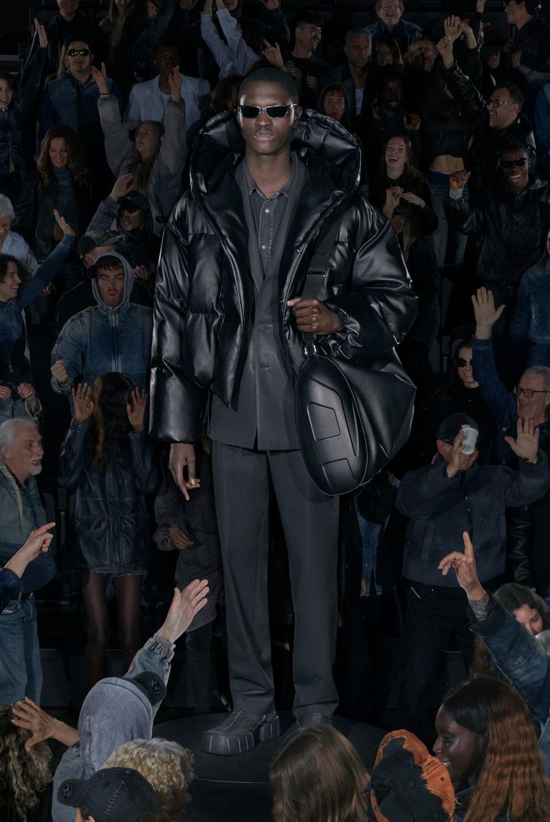 Fashion model wearing an oversized black puffer jacket and sunglasses stands on a runway with an audience in the background.