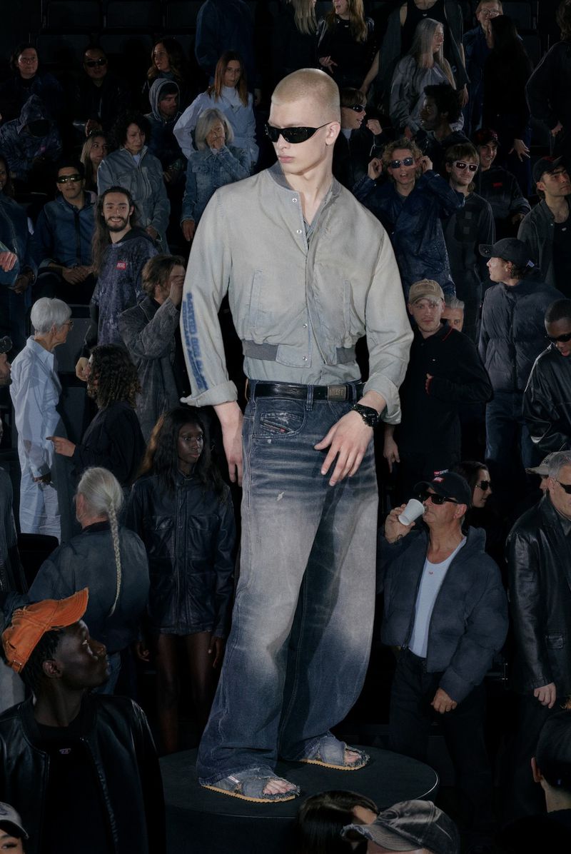 Male fashion model walks runway in light grey shirt, denim jeans, and sunglasses, showcasing fashion for an audience at a fashion show.