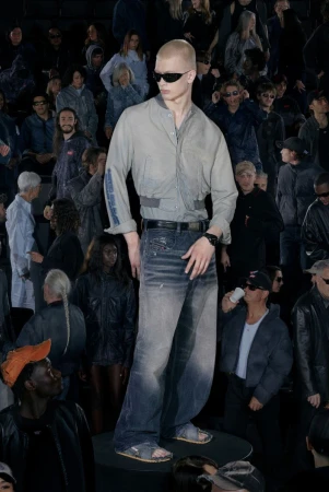 Male fashion model walks runway in light grey shirt, denim jeans, and sunglasses, showcasing fashion for an audience at a fashion show.