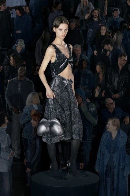 Runway fashion show featuring a model in a shimmering silver outfit carrying a unique purse, surrounded by a crowd of spectators.