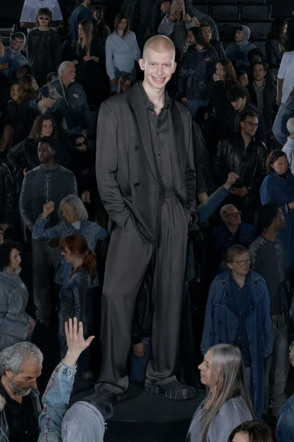 Male fashion model in a dark suit standing in front of a crowd. The model is smiling with a buzzcut. Fashion runway show or event.