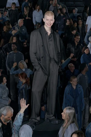 Male fashion model in a dark suit standing in front of a crowd. The model is smiling with a buzzcut. Fashion runway show or event.