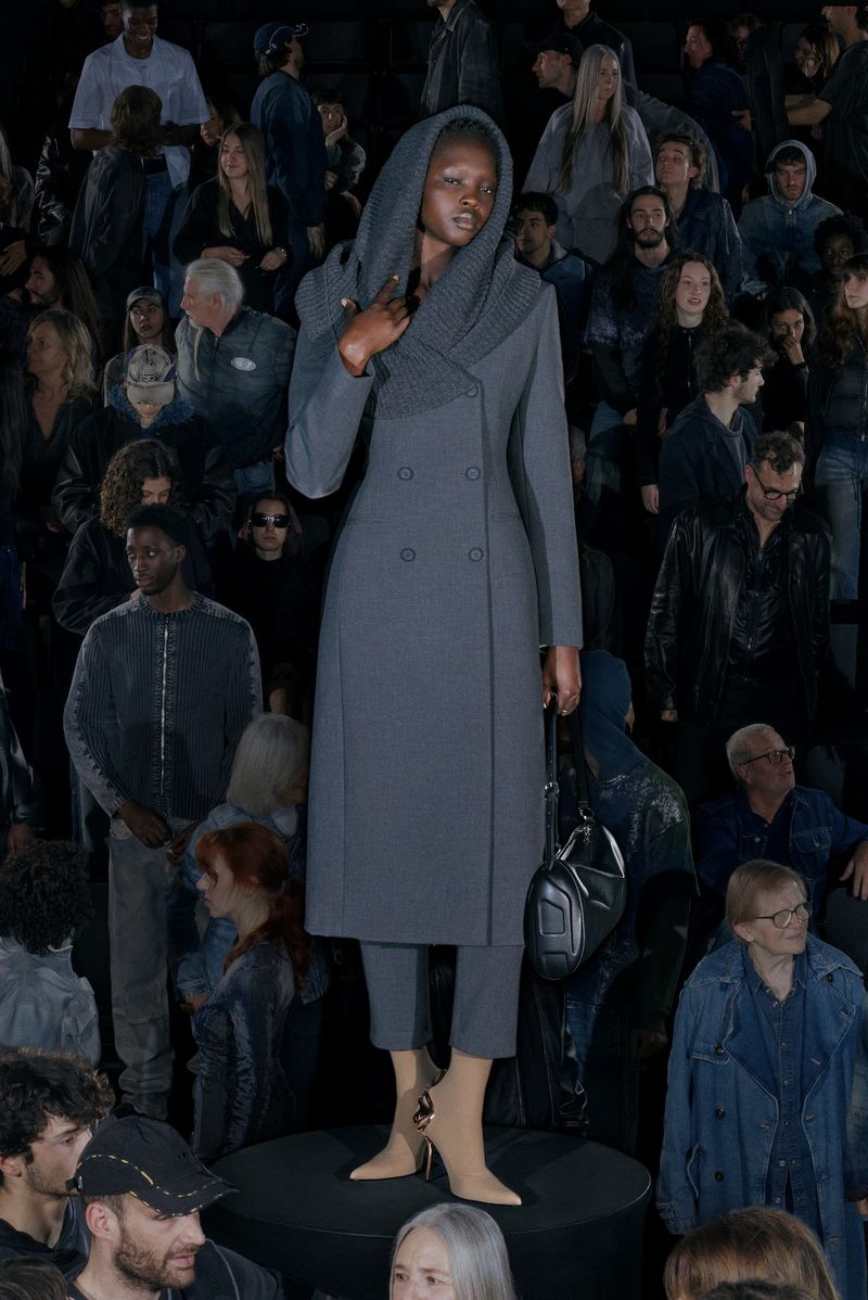 Runway fashion show with an African-descent female model in a gray double-breasted coat and beige heels, surrounded by a diverse audience.