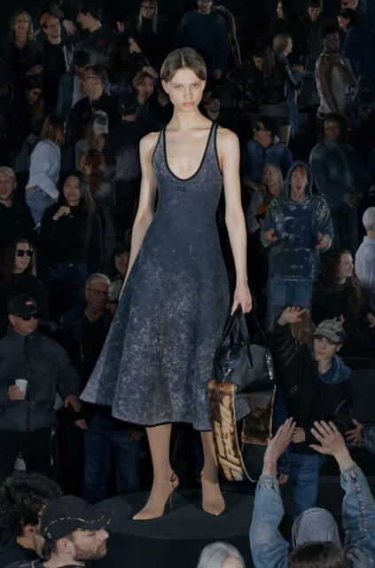 Fashion show features a model in a faded blue dress, carrying a black purse on the runway with a large crowd in the background.