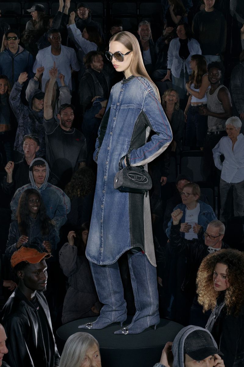 A runway shot with a woman in a long denim dress, sunglasses, and a purse, posing for the camera, surrounded by a crowd.