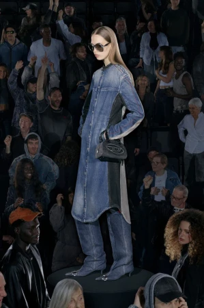 A runway shot with a woman in a long denim dress, sunglasses, and a purse, posing for the camera, surrounded by a crowd.