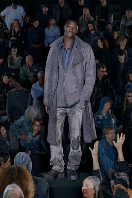 Runway fashion shot of a Black male model wearing a gray denim trench coat, jacket, and distressed jeans standing before a large crowd.