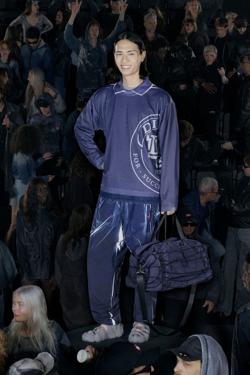 Full-length shot of a smiling Asian male model in a navy mesh top and sweatpants, posing with a duffel bag in front of a crowd.