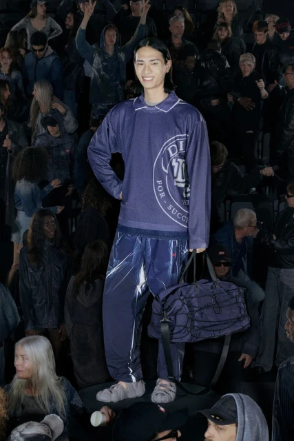 Full-length shot of a smiling Asian male model in a navy mesh top and sweatpants, posing with a duffel bag in front of a crowd.