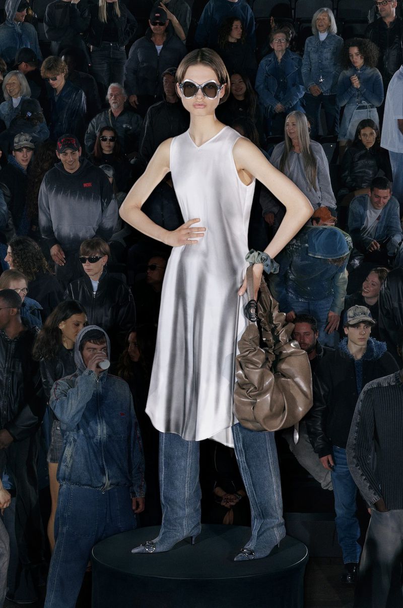 Fashion shot of a female model in a white dress and jeans, standing on a podium surrounded by a large group of people.