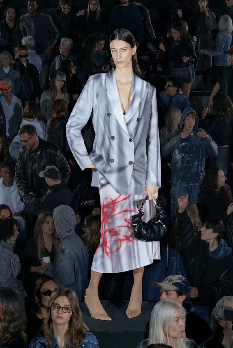 Runway fashion show featuring a woman in a gray and white blazer and skirt, carrying a black purse, surrounded by a crowd of spectators.