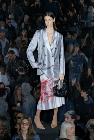 Runway fashion show featuring a woman in a gray and white blazer and skirt, carrying a black purse, surrounded by a crowd of spectators.