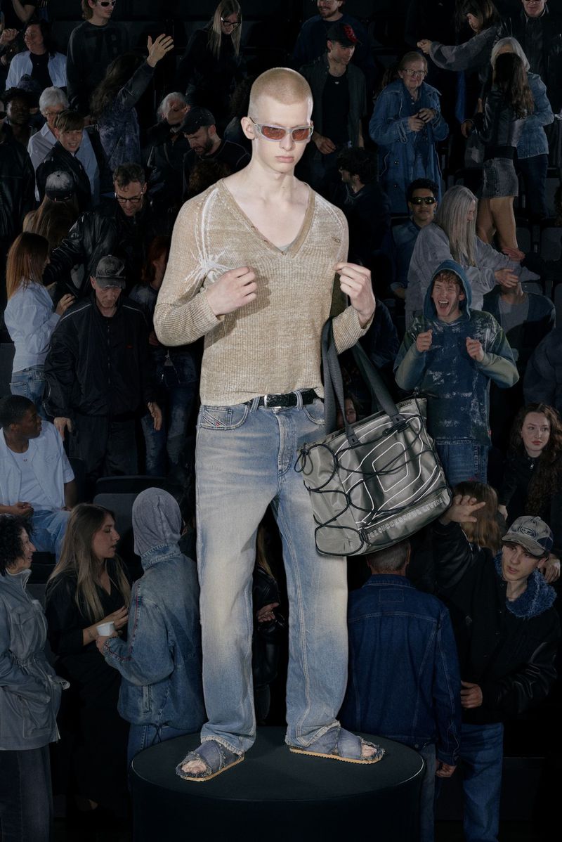 A fashion model poses on a black platform in light blue jeans, a tan V-neck sweater, sandals, and sunglasses in front of a crowd.