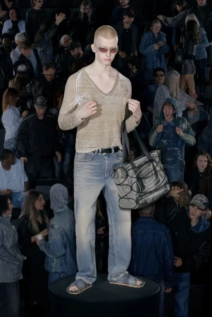 A fashion model poses on a black platform in light blue jeans, a tan V-neck sweater, sandals, and sunglasses in front of a crowd.