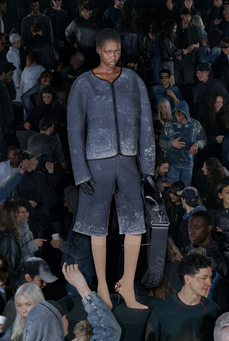 Fashion show featuring a Black model in a gray suit and nude tights, surrounded by a crowd in a dimly lit indoor venue.