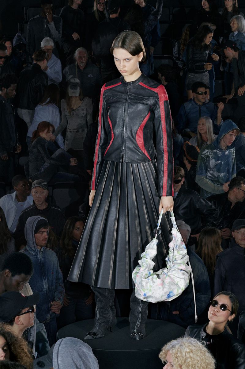 A model in a black leather outfit on a dark runway, holding a bag, surrounded by a blurred crowd in an indoor setting during a fashion show.